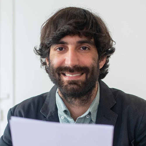 A caucasian, bearded man in mid-30s, at his desk, smiling casually at camera.